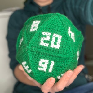 May include: A green crocheted dice with white numbers. The dice is being held in someone's hand.
