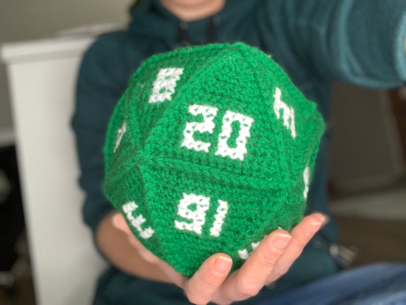 May include: A green crocheted dice with white numbers. The dice is being held in someone's hand.