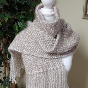 May include: A beige and white knitted scarf with a fringed edge. The scarf is displayed on a white mannequin head and has a button closure. The scarf appears to be handmade and is likely for cold weather.