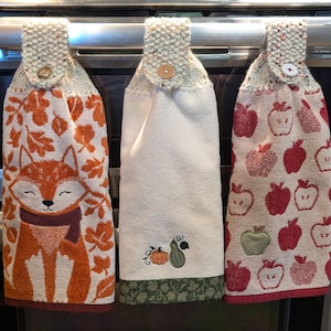May include: Three hanging kitchen towels with buttoned tops. The left towel features an orange fox and autumn leaves. The center towel is cream with a small pumpkin and gourd embroidery. The right towel displays a pattern of red and green apples.