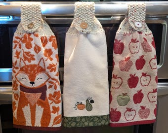 May include: Three hanging kitchen towels with buttoned tops. The left towel features an orange fox and autumn leaves. The center towel is cream with a small pumpkin and gourd embroidery. The right towel displays a pattern of red and green apples.