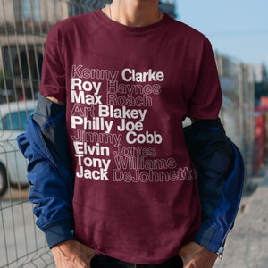 May include: A maroon t-shirt with white text listing the names of famous jazz drummers: Kenny Clarke, Roy Haynes, Max Roach, Art Blakey, Philly Joe, Jimmy Cobb, Elvin Jones, Tony Williams, and Jack DeJohnette.