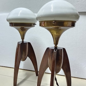 Może przedstawiać: Para lamp stołowych w stylu mid-century modern z białymi kloszami szklanymi i elementami mosiężnymi. Lampy mają unikalną podstawę trójnożną wykonaną z ciemnego drewna.