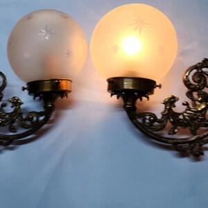 May include: A pair of antique brass wall sconces with frosted glass shades featuring a star pattern. The sconces have intricate, ornate designs.