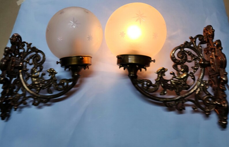 May include: A pair of antique brass wall sconces with frosted glass shades featuring a star pattern. The sconces have intricate, ornate designs.