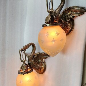 May include: Two brass swan wall sconces with frosted glass shades featuring a star pattern. The sconces are lit and the light shines through the glass.