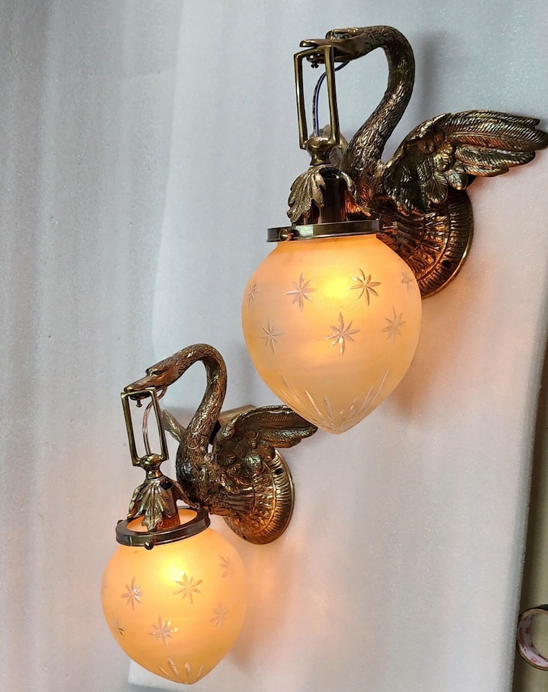 May include: Two brass swan wall sconces with frosted glass shades featuring a star pattern. The sconces are lit and the light shines through the glass.