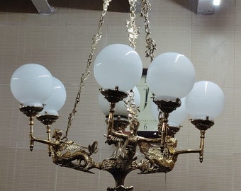 May include: A gold-toned chandelier with six white glass globes and a decorative design featuring mermaids. The chandelier is suspended from the ceiling by a chain.