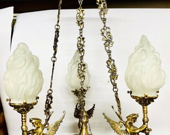 May include: Antique brass chandelier with three frosted glass shades shaped like flames. The chandelier features ornate details, including winged figures and decorative chains. A vintage lighting fixture.
