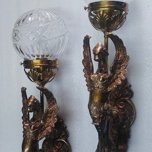 May include: Pair of antique wall sconces. Each sconce features a clear, cut-glass globe shade atop a brass fixture. The brass fixtures are adorned with a winged figure design. The sconces are mounted on a white wall.