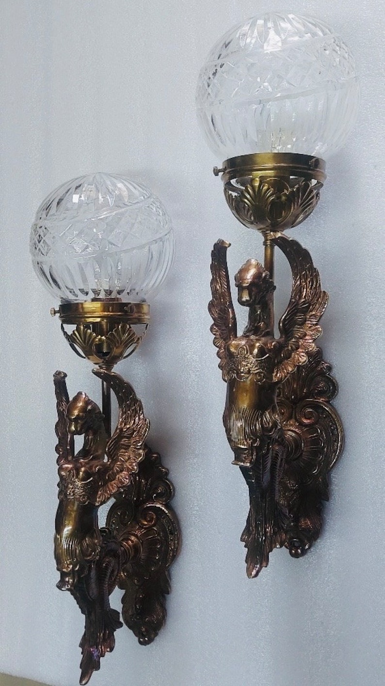 May include: Pair of antique wall sconces. Each sconce features a clear, cut-glass globe shade atop a brass fixture. The brass fixtures are adorned with a winged figure design. The sconces are mounted on a white wall.