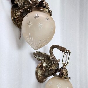 May include: Two antique brass swan wall sconces with frosted glass shades featuring a starburst pattern. The sconces have a vintage aesthetic and would add a touch of elegance to any room.