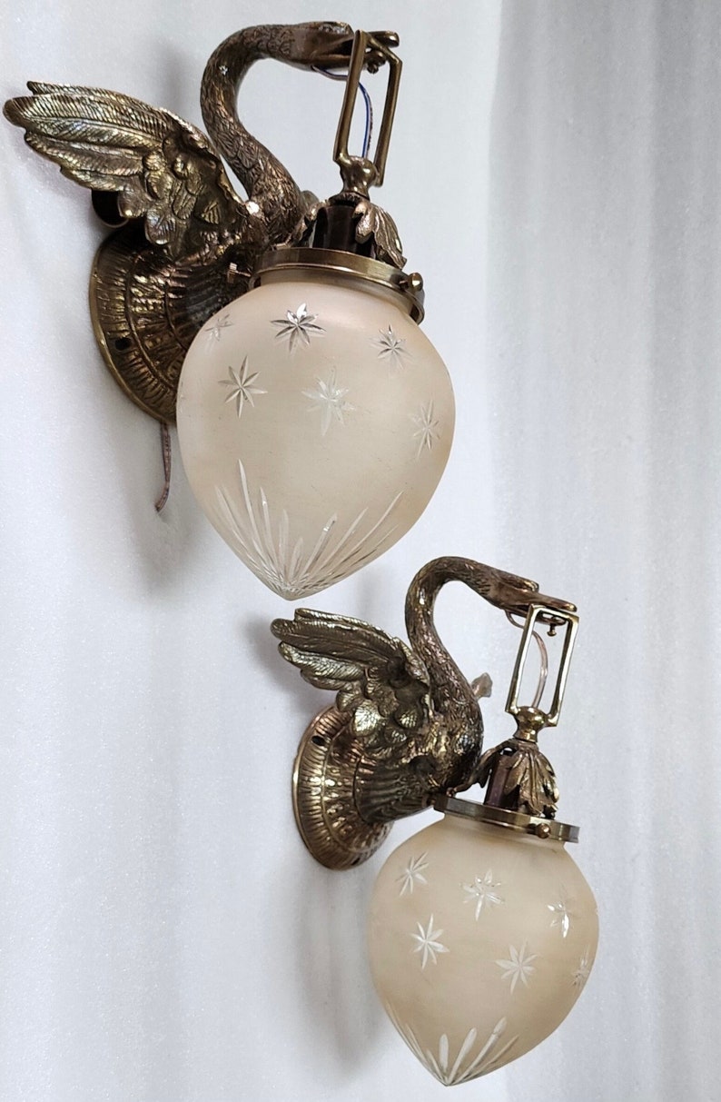 May include: Two antique brass swan wall sconces with frosted glass shades featuring a starburst pattern. The sconces have a vintage aesthetic and would add a touch of elegance to any room.