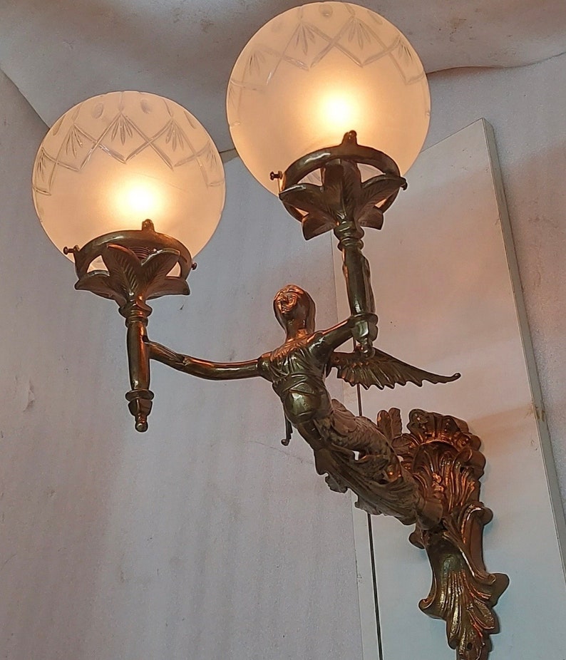 May include: A brass wall sconce with a winged figure holding two frosted glass globes. The globes have a geometric pattern and are lit from within.