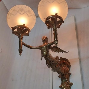 May include: A gold-toned wall sconce with a winged figure holding two frosted glass globes. The sconce has a decorative scrollwork design.