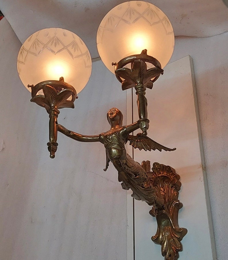 May include: A gold-toned wall sconce with a winged figure holding two frosted glass globes. The sconce has a decorative scrollwork design.