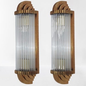 Op de afbeelding: Twee wandkandelaars in Art Deco-stijl. Elke kandelaar heeft een verticale opstelling van heldere, gegroefde glazen staven in een rechthoekig frame. De frames zijn warm goudbruin van kleur met decoratieve messing accenten.