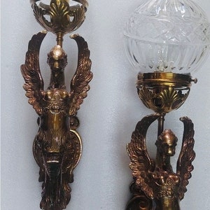 May include: Pair of antique wall sconces featuring a winged figure design. Each sconce has a clear, globe-shaped glass shade and a bronze-colored metal frame. The winged figure is detailed with intricate feather and body details.