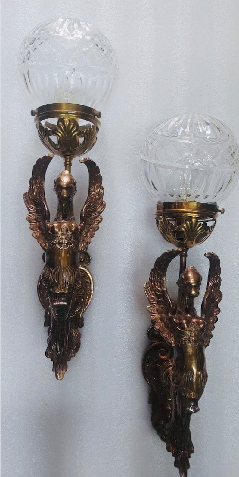 May include: Pair of antique wall sconces featuring a winged figure design. Each sconce has a clear, globe-shaped glass shade and a bronze-colored metal frame. The winged figure is detailed with intricate feather and body details.