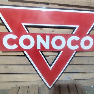 May include: Vintage Conoco gas station sign. The sign features a red triangle with a white border and the word "CONOCO" in white block letters. The sign is mounted on a wooden pallet background.