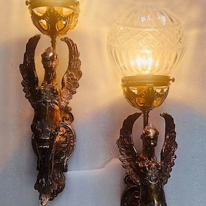 May include: Pair of antique wall sconces with a bronze finish, featuring winged figures supporting a clear, globe-shaped glass shade. The sconces are illuminated, casting a warm glow. Decorative lighting for home decor.