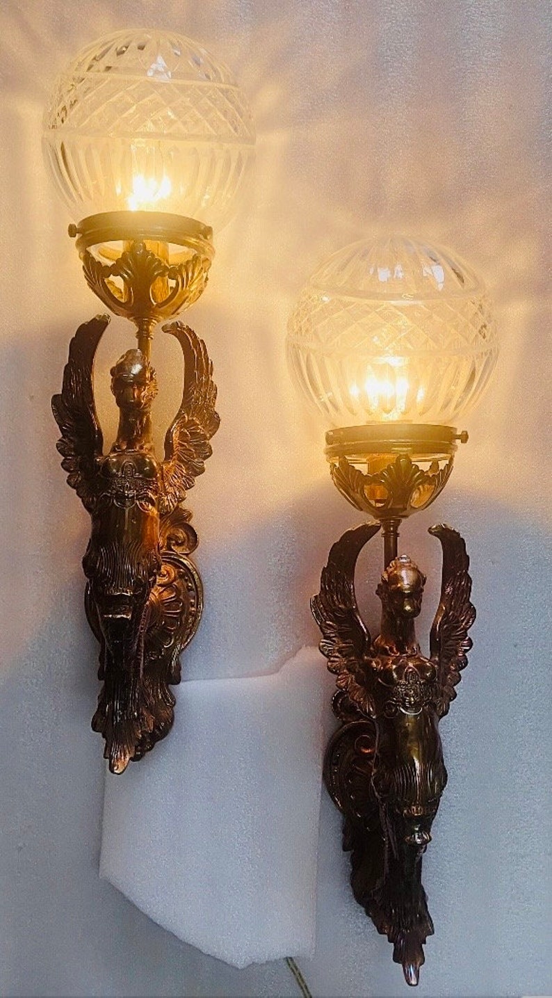 May include: Pair of antique wall sconces with a bronze finish, featuring winged figures supporting a clear, globe-shaped glass shade. The sconces are illuminated, casting a warm glow. Decorative lighting for home decor.