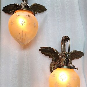 May include: Two brass wall sconces with frosted glass shades featuring a starburst design. Each sconce is shaped like a bird with outstretched wings.