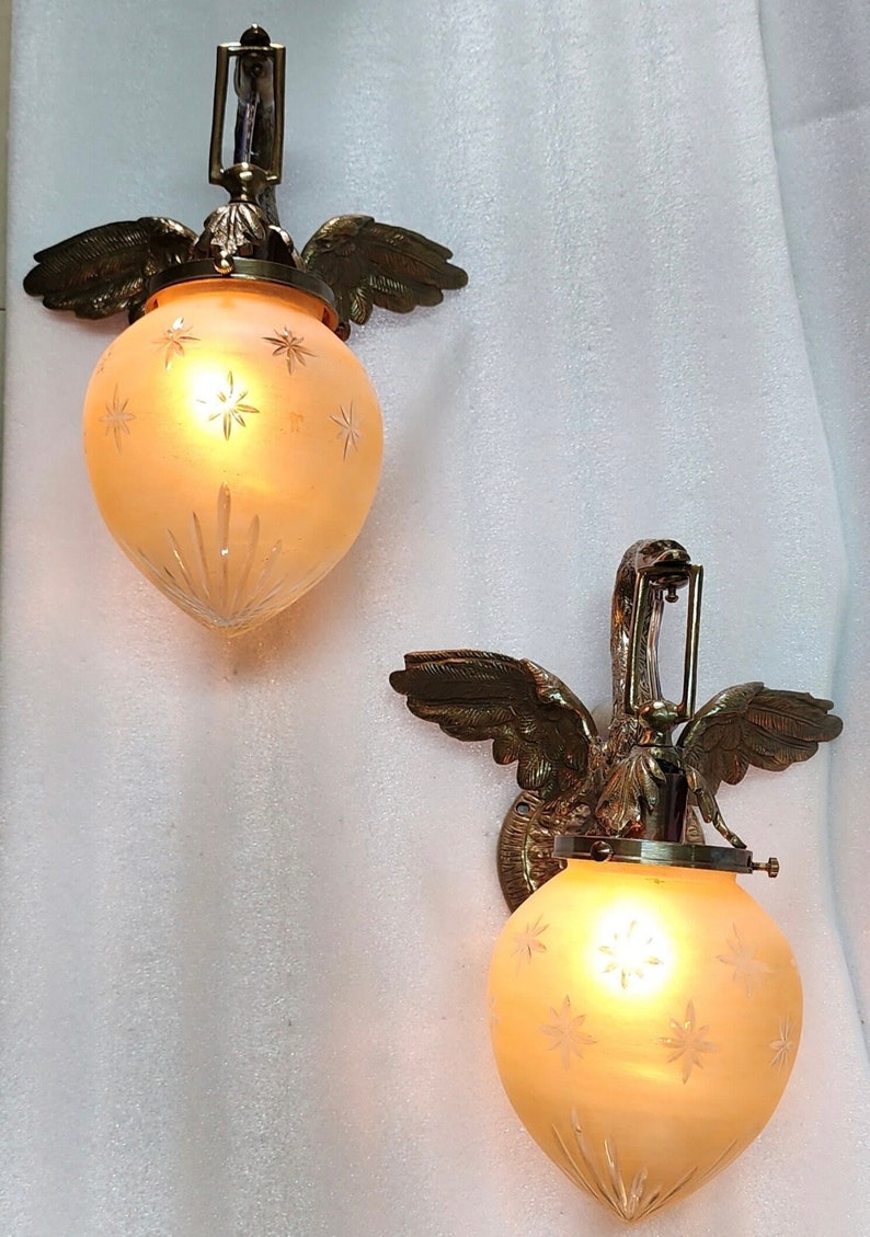 May include: Two brass wall sconces with frosted glass shades featuring a starburst design. Each sconce is shaped like a bird with outstretched wings.