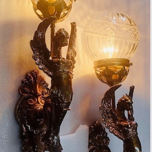 May include: Two antique wall sconces with a bronze finish. Each sconce features a winged figure holding a clear, globe-shaped glass shade. The shades have a cut-glass design, and the sconces are mounted on a wall.