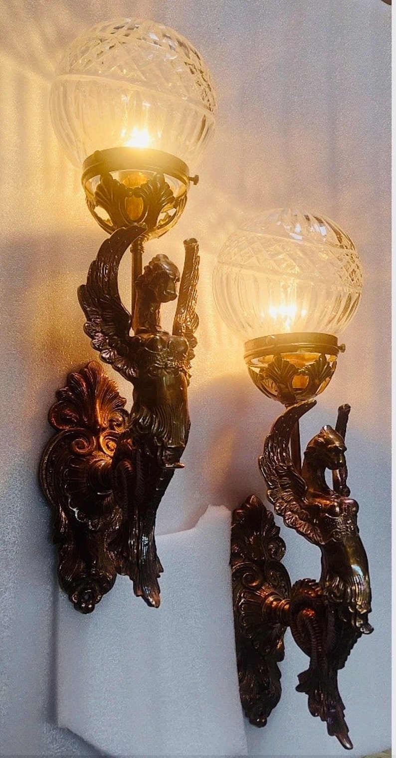 May include: Two antique wall sconces with a bronze finish. Each sconce features a winged figure holding a clear, globe-shaped glass shade. The shades have a cut-glass design, and the sconces are mounted on a wall.