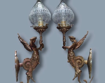Könnte beinhalten: Ein Paar antike Wandleuchter aus Messing mit gravierten Glasschirmen. Jeder Leuchter zeigt eine geflügelte Figur, die einen Kerzenhalter hält.
