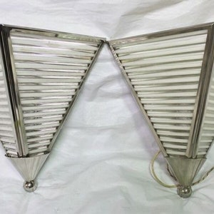 May include: A pair of silver metal wall sconces with a geometric design. Each sconce has a triangular shape with a series of horizontal slats.