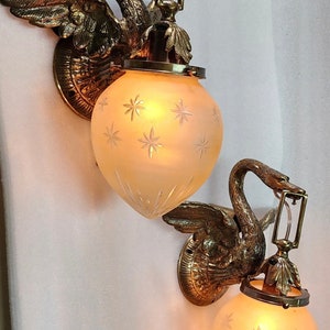 May include: A pair of antique brass swan wall sconces with frosted glass shades featuring a starburst pattern. The sconces are lit and casting a warm glow.