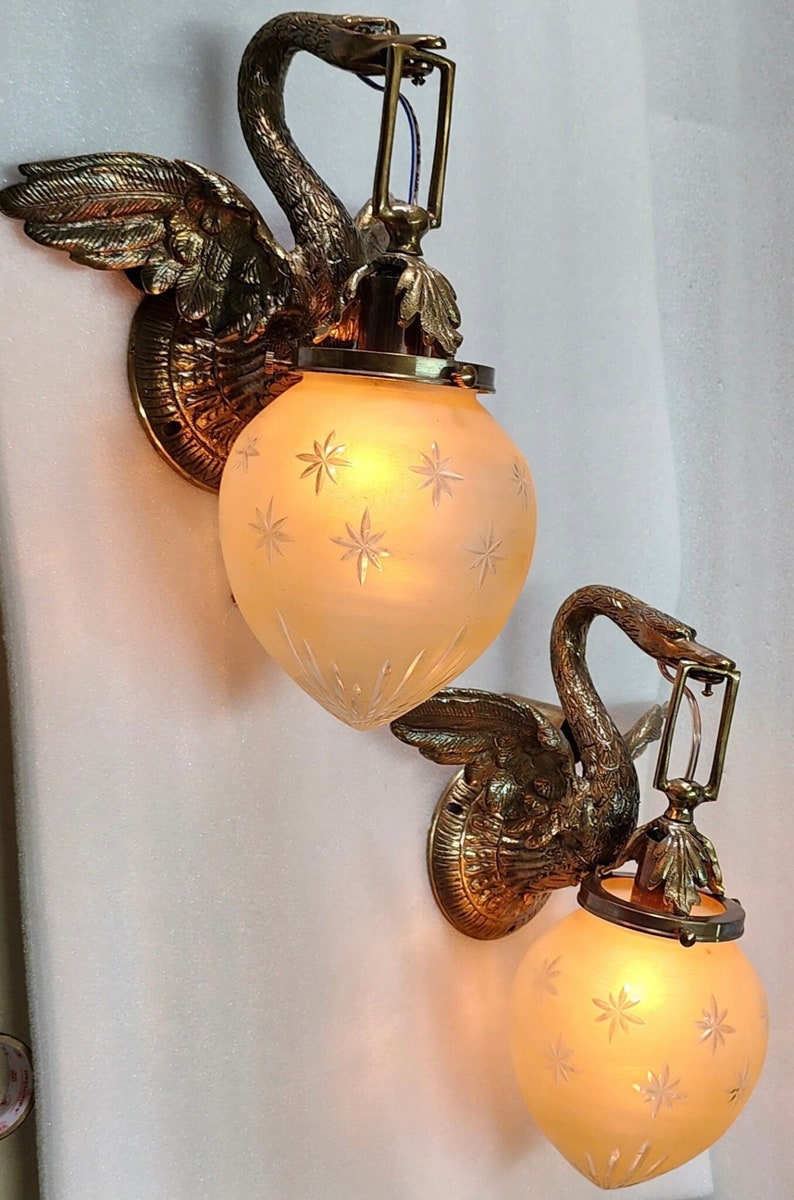 May include: A pair of antique brass swan wall sconces with frosted glass shades featuring a starburst pattern. The sconces are lit and casting a warm glow.