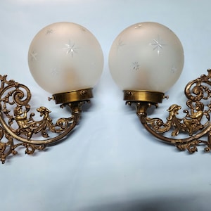 May include: A pair of antique brass wall sconces with frosted glass globes featuring a star pattern. Each sconce has intricate scrollwork and a cherub figure.