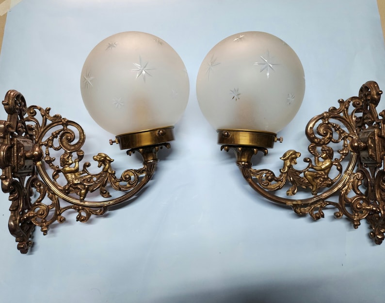 May include: A pair of antique brass wall sconces with frosted glass globes featuring a star pattern. Each sconce has intricate scrollwork and a cherub figure.