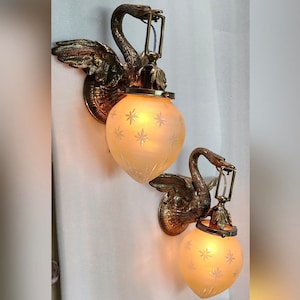 May include: Two antique brass swan wall sconces with frosted glass shades featuring a starburst pattern. The sconces have a vintage aesthetic and would add a touch of elegance to any room.