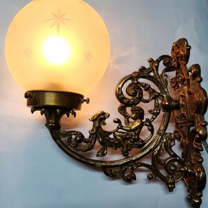 May include: A brass wall sconce with an ornate design featuring a cherub figure. The sconce has a frosted glass globe shade.
