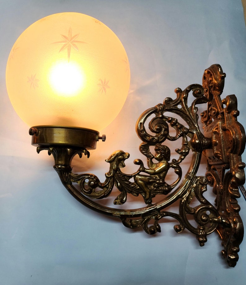 May include: A brass wall sconce with an ornate design featuring a cherub figure. The sconce has a frosted glass globe shade.