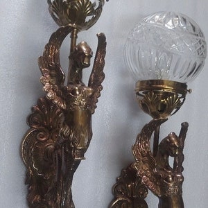 May include: Pair of antique wall sconces with a bronze finish. Each sconce features a winged figure holding a clear, globe-shaped glass shade. The glass shades have a textured, cut-glass design. Ornate, decorative details are present.