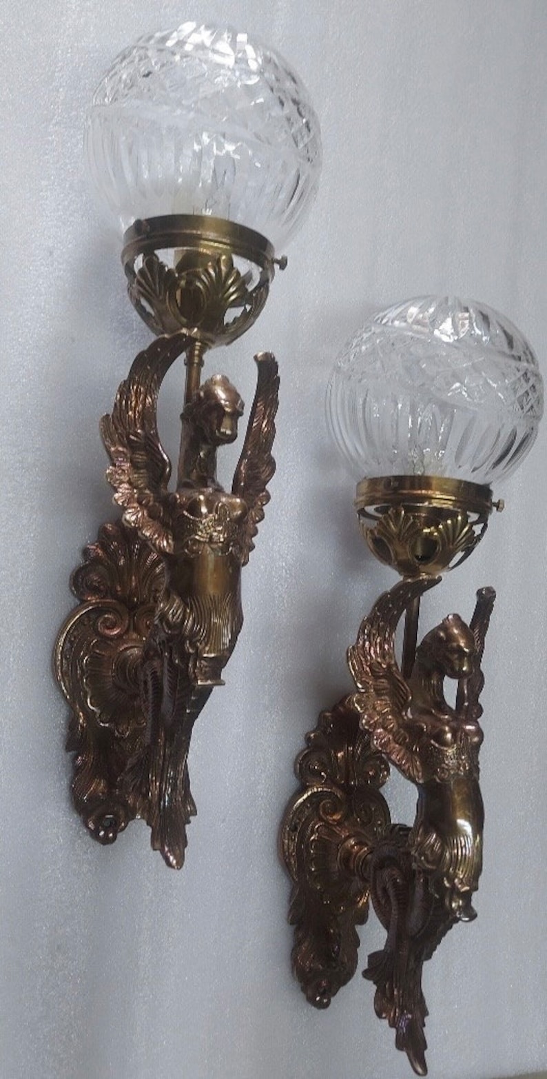 May include: Pair of antique wall sconces with a bronze finish. Each sconce features a winged figure holding a clear, globe-shaped glass shade. The glass shades have a textured, cut-glass design. Ornate, decorative details are present.