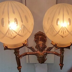 May include: A brass wall sconce with two frosted glass globes, each with a decorative etched pattern. The sconce features a central figure of a woman with wings.