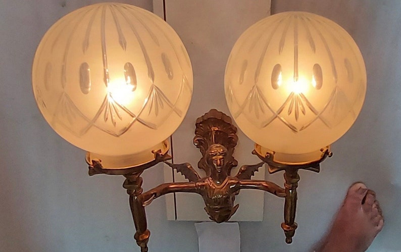 May include: A brass wall sconce with two frosted glass globes, each with a decorative etched pattern. The sconce features a central figure of a woman with wings.