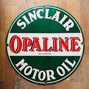 May include: A round, green and white metal sign with the text "Sinclair Opaline Motor Oil" in white and red lettering. The text "Reg. U.S. Pat. Off." is printed in black below the word "Opaline".