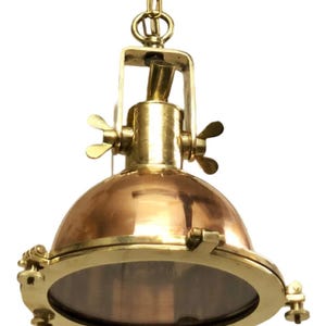 May include: A brass and copper nautical pendant light fixture with a clear glass lens. The light has a chain for hanging and adjustable side brackets.