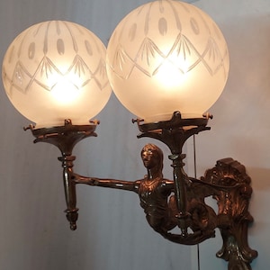 May include: A bronze wall sconce with two frosted glass globes and a decorative figure of a woman holding a torch. The globes have a diamond pattern etched into the glass.