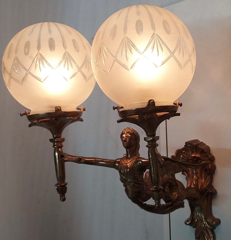 May include: A bronze wall sconce with two frosted glass globes and a decorative figure of a woman holding a torch. The globes have a diamond pattern etched into the glass.
