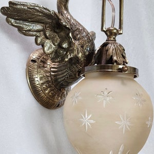 May include: A brass swan wall sconce with a frosted glass shade featuring a starburst pattern. The sconce has a decorative base and a simple, elegant design.