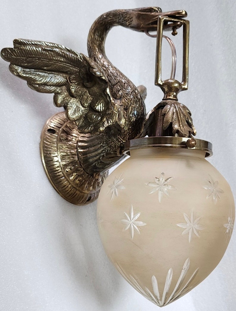 May include: A brass swan wall sconce with a frosted glass shade featuring a starburst pattern. The sconce has a decorative base and a simple, elegant design.