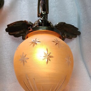 May include: A frosted glass pendant light fixture with a brass finish and a starburst pattern. The light fixture is suspended from a chain and has a decorative winged design.
